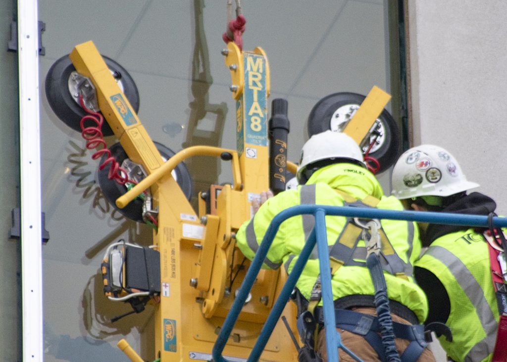 Two installers using an MRT‑A8 vacuum lifter to position a large glass panel on a building façade.